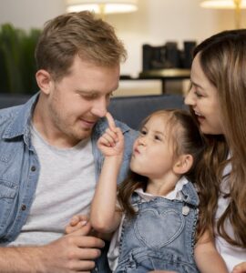 Uomo e donna seduti su un divano con una bambina tra loro. La bambina tocca il naso dell'uomo con il dito mentre la donna la osserva sorridendo. Scena di interazione familiare in ambiente domestico.