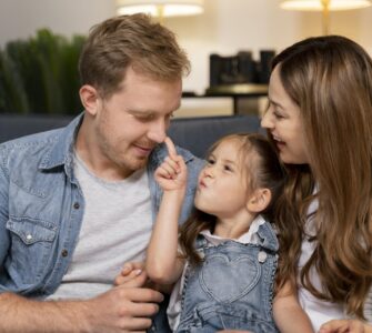 Uomo e donna seduti su un divano con una bambina tra loro. La bambina tocca il naso dell'uomo con il dito mentre la donna la osserva sorridendo. Scena di interazione familiare in ambiente domestico.