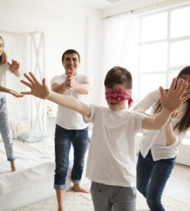 Una famiglia di quattro persone gioca allegramente a mosca cieca in una camera da letto luminosa. Al centro, un ragazzo bendato protende le braccia, mentre i genitori e la sorella gli si muovono intorno sorridendo per non farsi acchiappare.