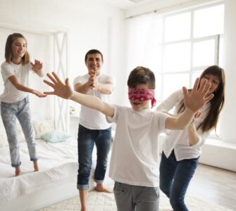 Una famiglia di quattro persone gioca allegramente a mosca cieca in una camera da letto luminosa. Al centro, un ragazzo bendato protende le braccia, mentre i genitori e la sorella gli si muovono intorno sorridendo per non farsi acchiappare.