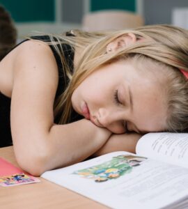 Bambina annoiata in classe che dorme con la testa poggiata sul banco e un libro aperto davanti.