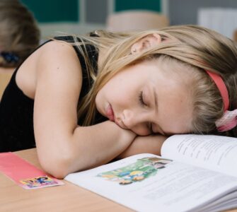 Bambina annoiata in classe che dorme con la testa poggiata sul banco e un libro aperto davanti.