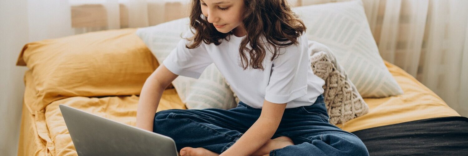 bambina che studia a casa seduta sul letto con davanti un computer e un quaderno con poggiata su una penna