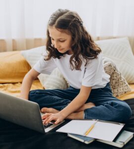 bambina che studia a casa seduta sul letto con davanti un computer e un quaderno con poggiata su una penna