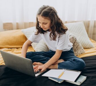 bambina che studia a casa seduta sul letto con davanti un computer e un quaderno con poggiata su una penna