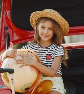 Una bambina seduta nel bagagliaio aperto di un’auto rossa, sorridente, con indosso un cappello di paglia, una maglietta a righe bianche e nere e pantaloncini chiari. Tiene in braccio un grande mappamondo e fa il gesto del pollice in su. Accanto a lei ci sono una valigia con manico estensibile e una borsa colorata a righe. La scena si svolge all’aperto, in un contesto estivo.