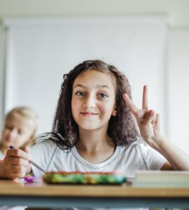 Una bambina con i capelli ricci e scuri sorride seduta a un banco di scuola, all'interno di un'aula luminosa, facendo il segno di vittoria con la mano sinistra.