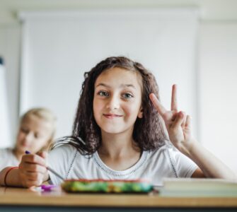Una bambina con i capelli ricci e scuri sorride seduta a un banco di scuola, all'interno di un'aula luminosa, facendo il segno di vittoria con la mano sinistra.